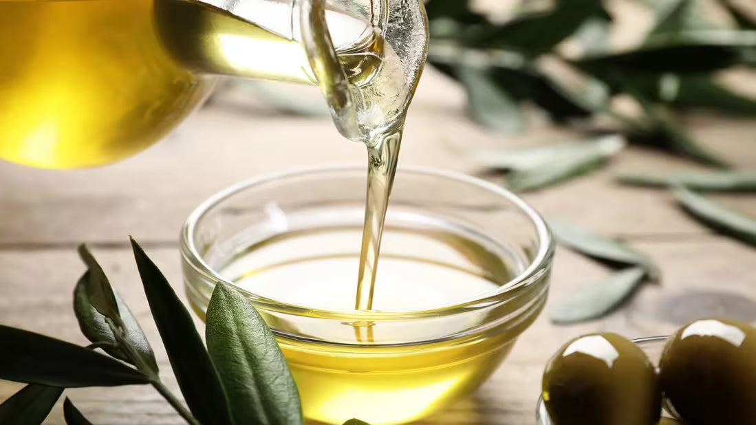 Olive oil being poured into a glass bowl with olive leaves on a wooden surface